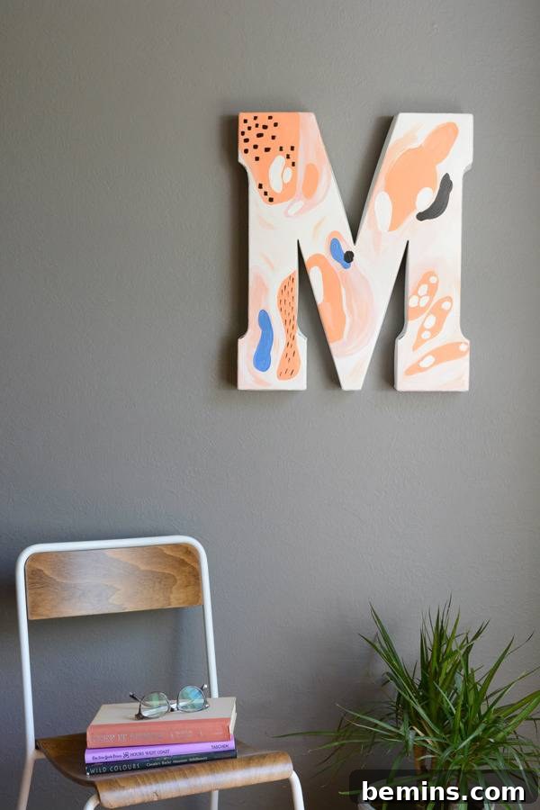 The abstract letter M brightens a cozy corner, hanging on a wall above a lush plant and a comfortable chair adorned with books, creating an inviting reading nock.