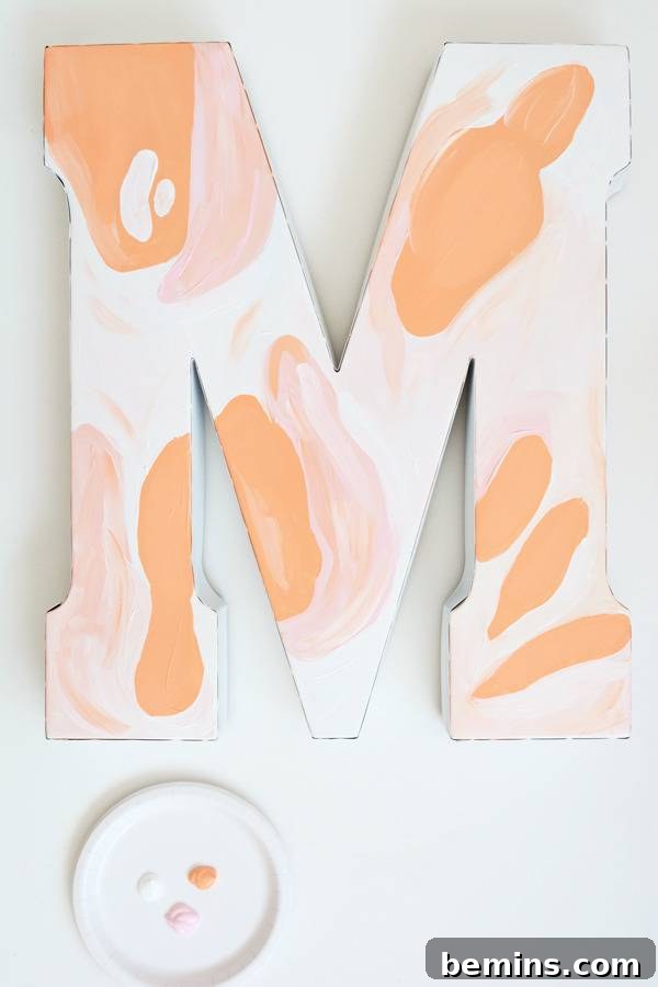 The partially painted letter M, featuring initial strokes of peach and pink, hangs on a light-colored wall, revealing the artistic process.