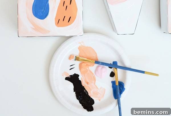 A paper plate holds vibrant blobs of black, orange, pink, and blue paint, alongside two paintbrushes, illustrating the color palette used for the abstract design.