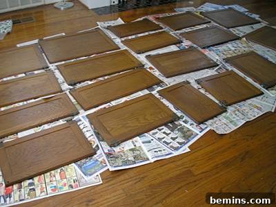 The front pieces of numerous wooden kitchen cabinets lay on newspaper spread out on a hardwoord floor, organized for painting.