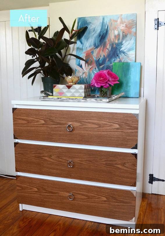 A brown and white three drawer dresser with a black plant, pink flowers and a blue abstract painting on top of it. - The completed dresser.