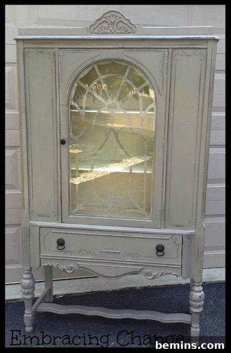 A white antique dining room hutch with a window on the front, standing gracefully in front of a white brick wall.
