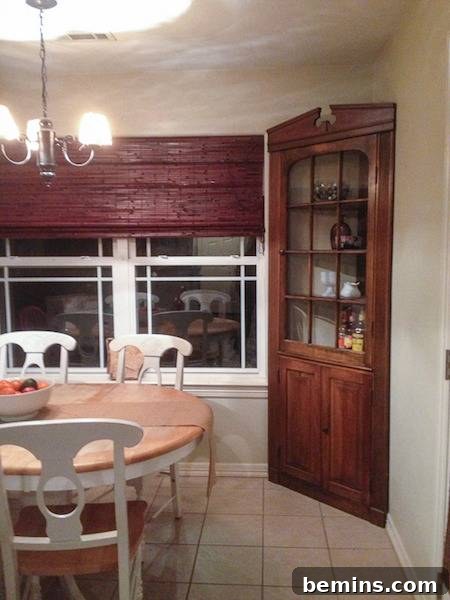 A dining room featuring a brown blind on the window and a brown display shelf tucked in the corner.