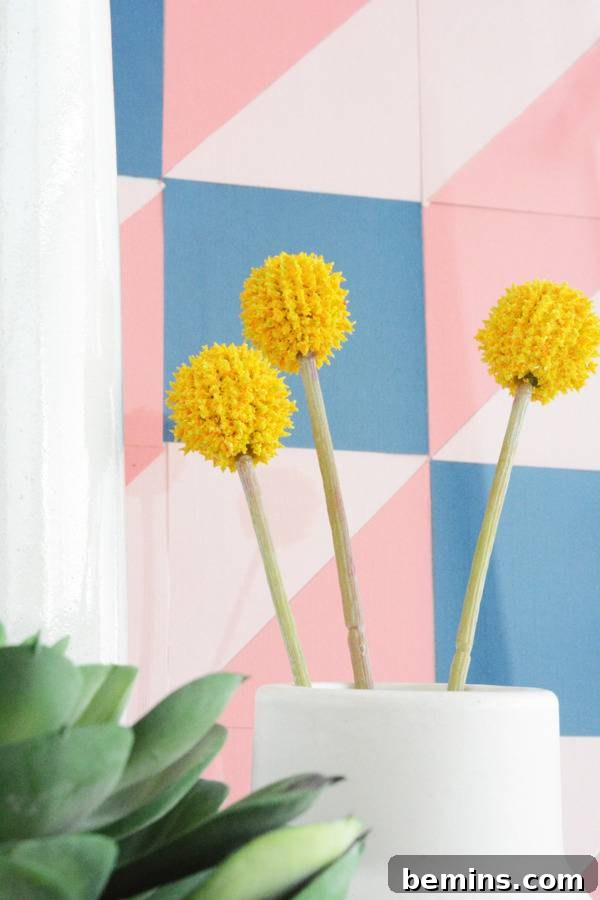 A flower with yellow balls is sitting in a cup near a colorful pastel wall.