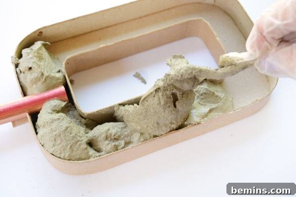 A hand dropping a scoop of freshly mixed cement into a rectangular cardboard mold, filling it carefully.