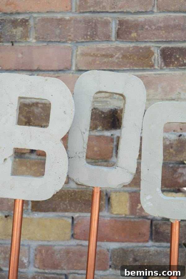 Three cut out concrete letters on copper rods spelling out the word "BOO," displayed elegantly in an outdoor setting.