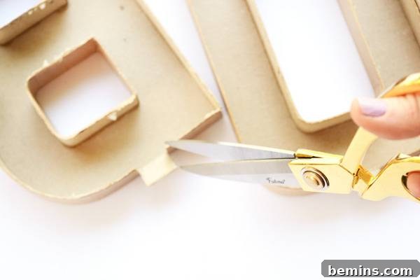 A pair of gold scissors poised to cut into the tan cardboard mold of a letter, creating a slot for the copper pipe.