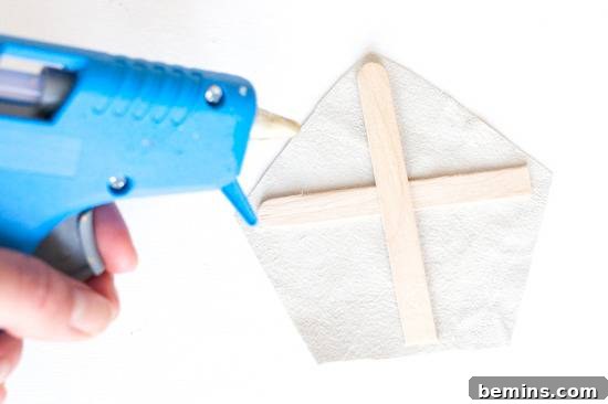 A woman's hand holding a blue glue gun, poised to glue popsicle sticks onto white paper.