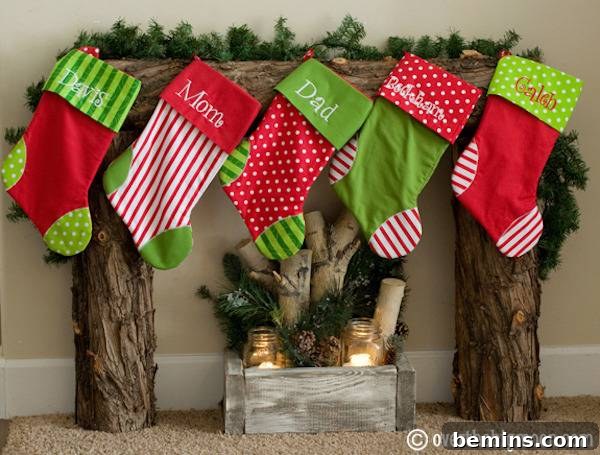 Five green, red and white stocking hanging from a tree trunk mantel over lit candles in a box of wood.