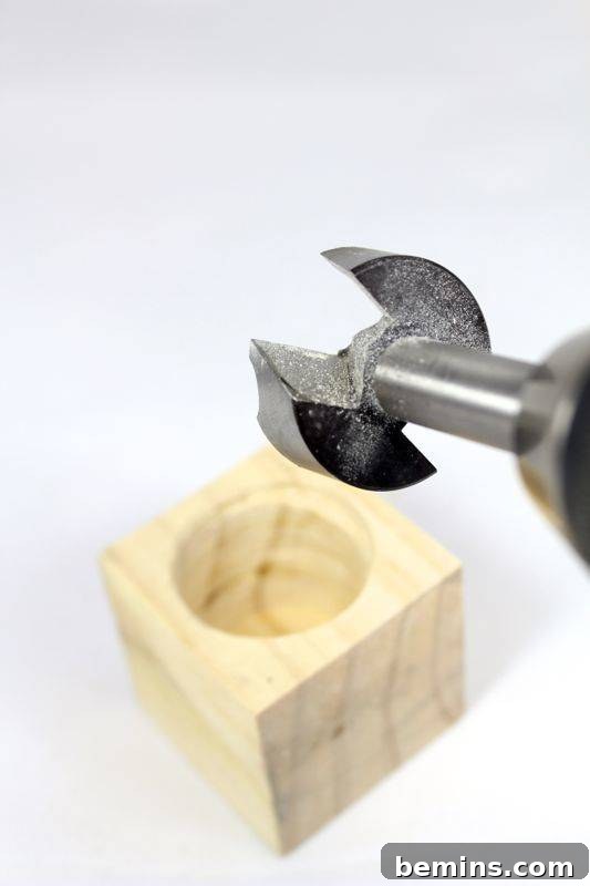 A drill bit is positioned above a light-colored wood block with a partially bored hole.