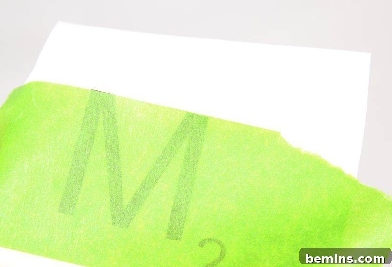The letter M is being carefully transferred onto a green surface with tape.