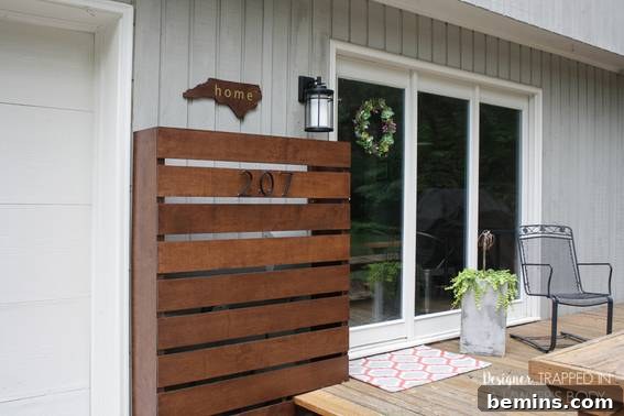 The exterior of a home with eight brown wooden slats next to three full sized windows and an orange and white door mat.