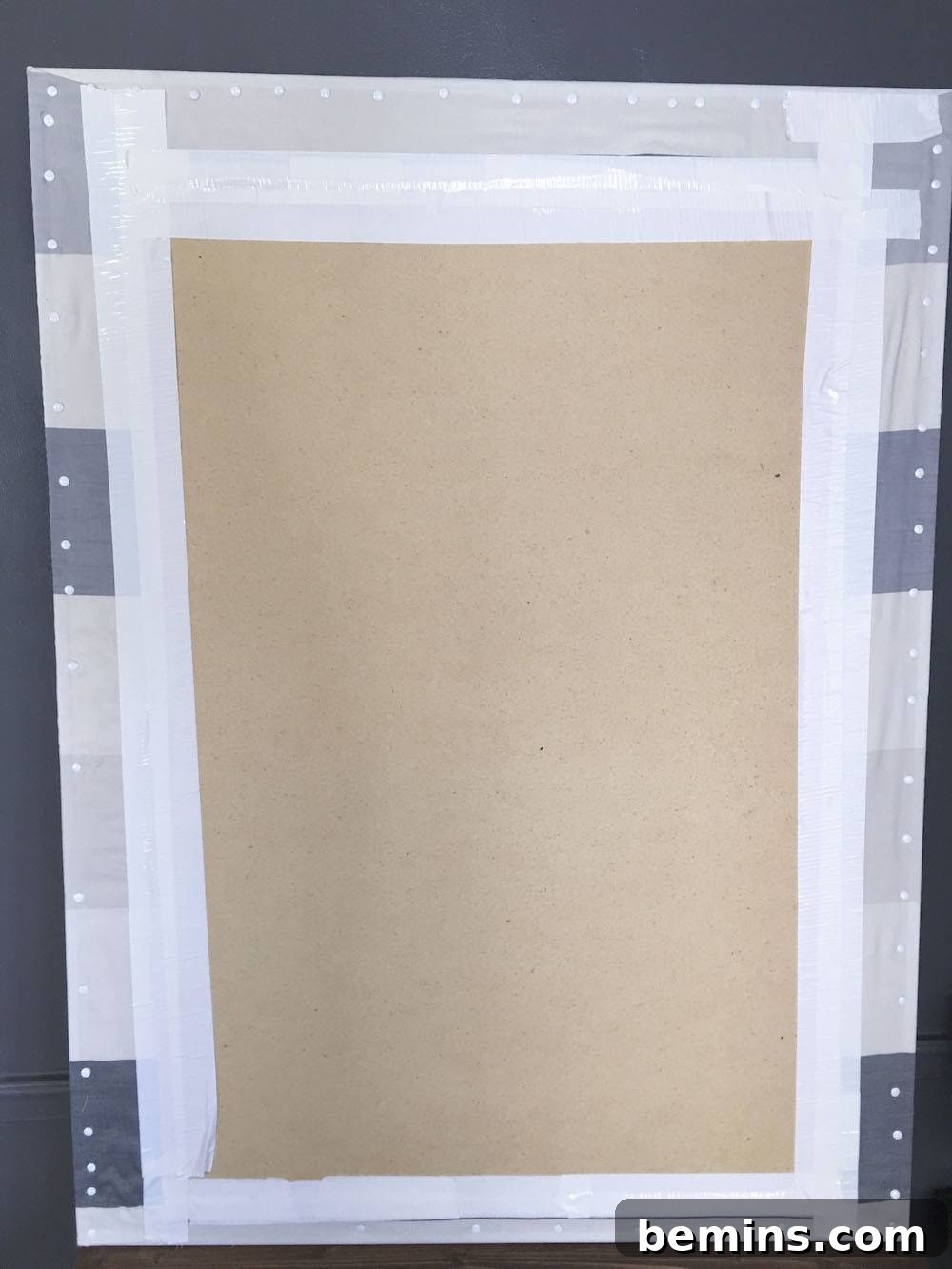 Securing the fabric to the back of the bulletin board with thumbtacks.