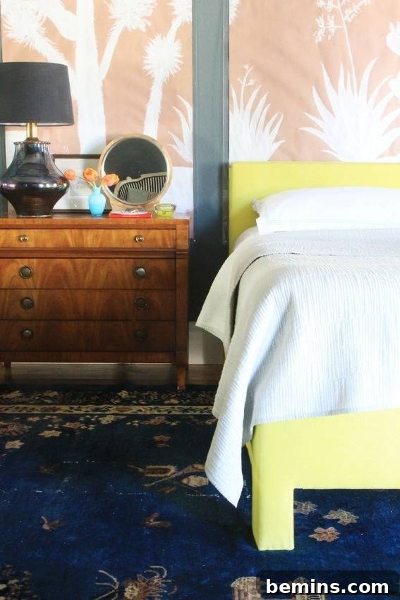 A yellow bed with white sheets next to a brown dresser that has a grey lamp on it.
