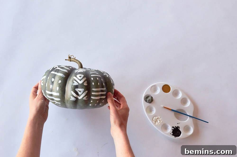 Two hands holding a pumpkin that has been painted grey and white next to a white painter's pallet.
